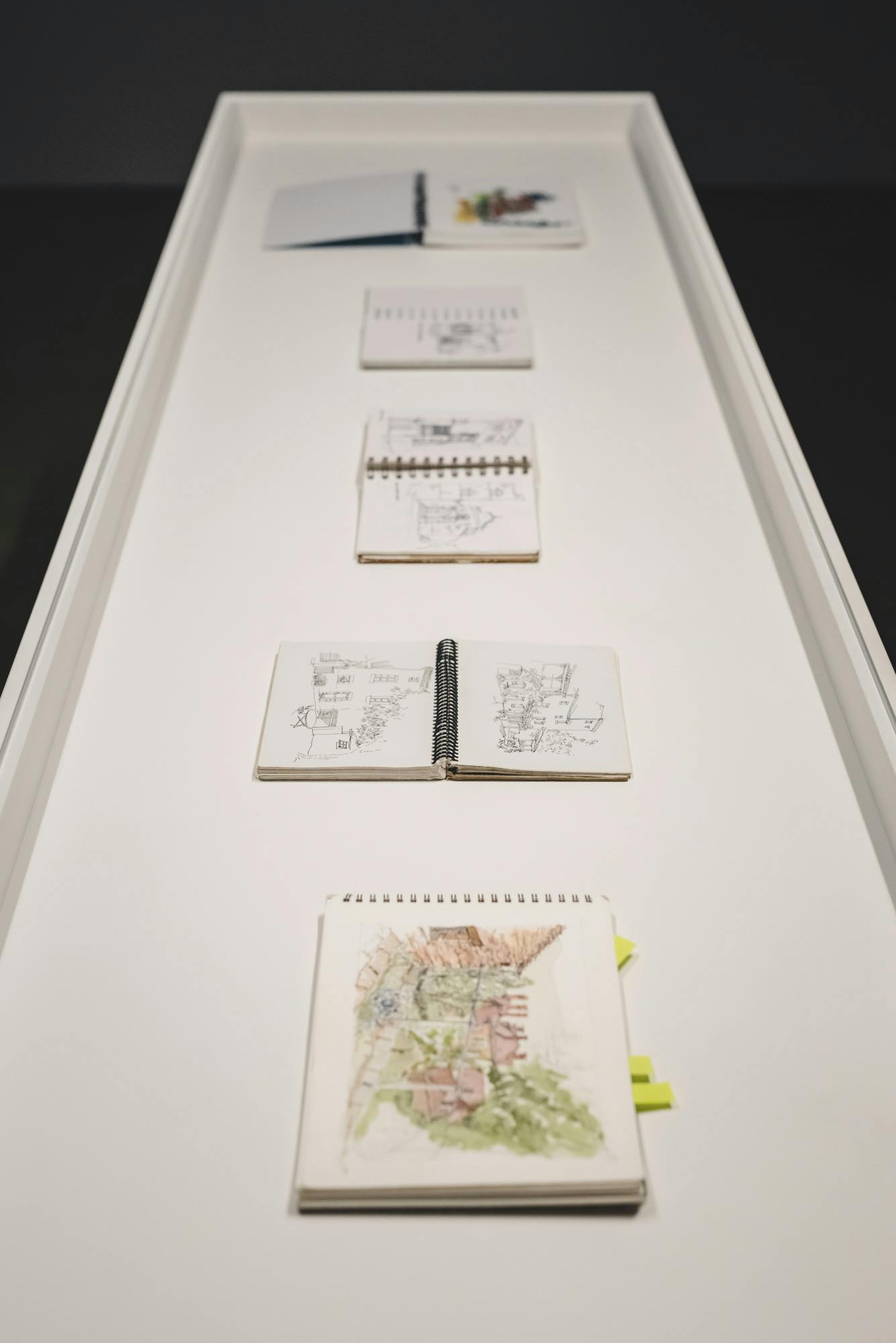 A white vitrine which contains five open sketchbooks showing images on buildings and landscapes.