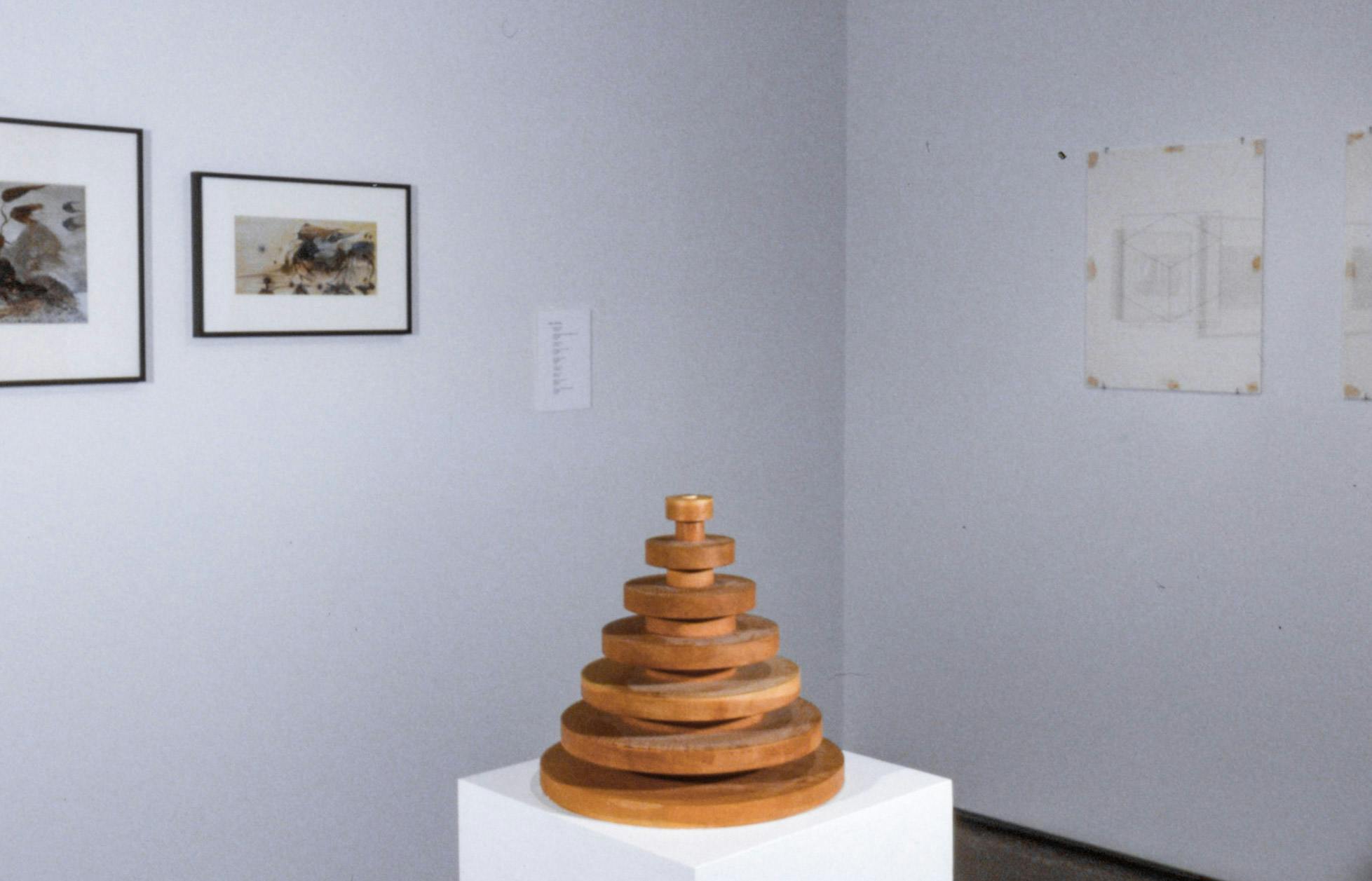 A coil-shaped wood sculpture is placed on a pedestal. Two coloured prints and a pair of black and white drawings are mounted on the wall behind this wood sculpture.  