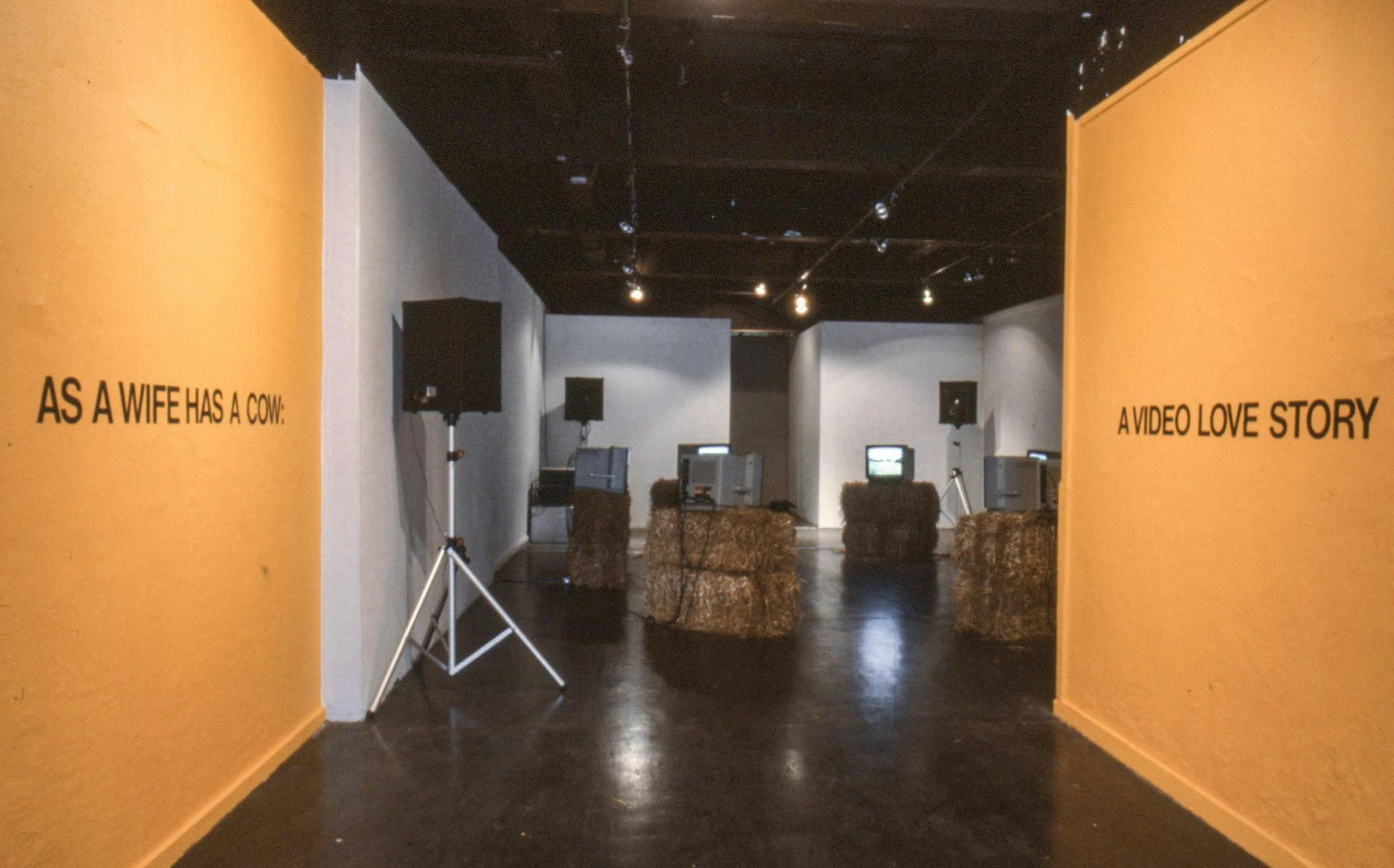 In a dimly lit gallery, there are 6 bales of hay with 6 TV's playing the same video. The entrance is painted yellow-orange, and black text on the walls reads "As A Wife Has A Cow: A Video Love Story."