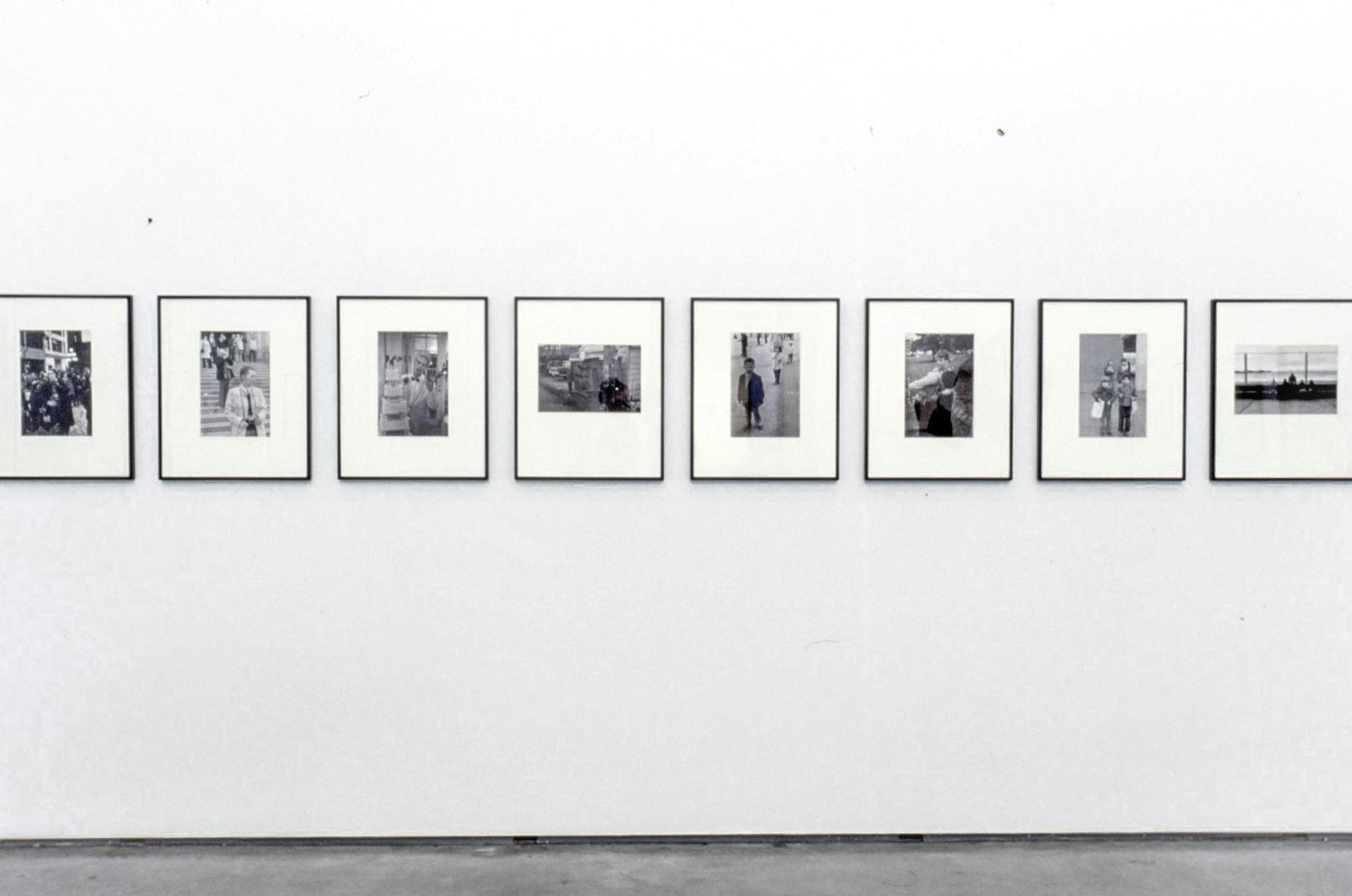 Eight black and white photographs are mounted on the wall. They are placed in a row horizontal to the floor. Many of those photographs depict pedestrians on the streets. 