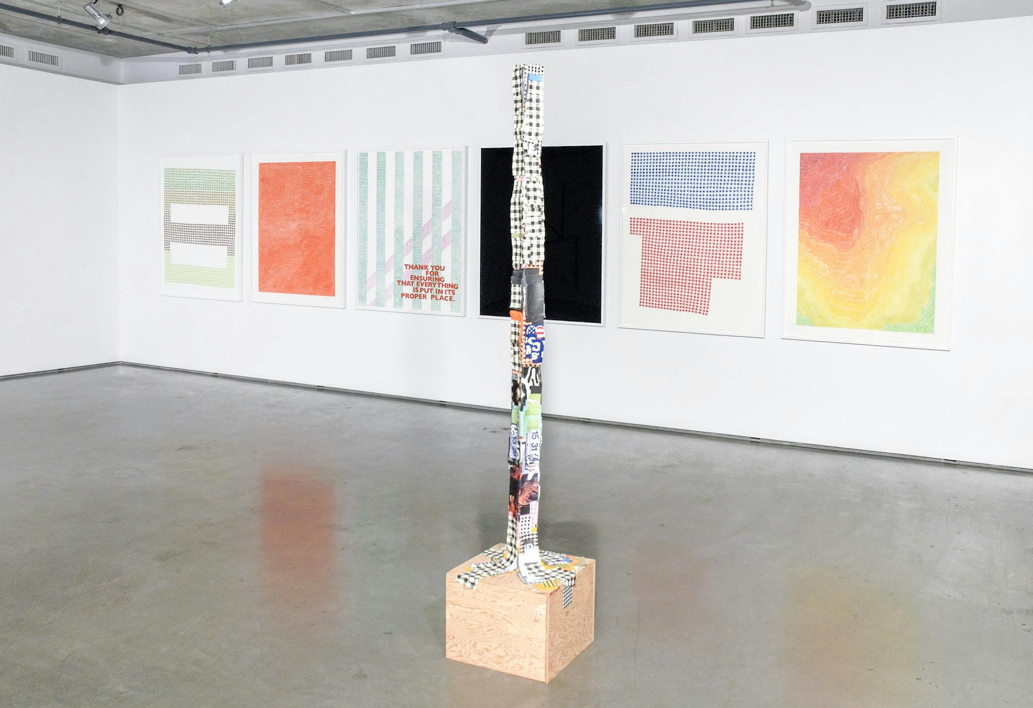 Various artworks are installed in a gallery space. Six large-sized two-dimensional works are mounted on the gallery walls. A thin tall sculpture covered with cutout papers stands on the floor.  