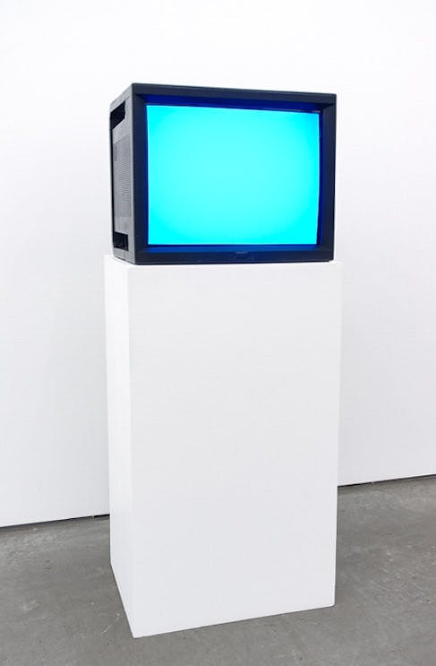 An installation view of a work titled Watercolour by Ceal Floyer. A CRT TV installed on a white pedestal displays a surface of a white screen illuminated in blue.