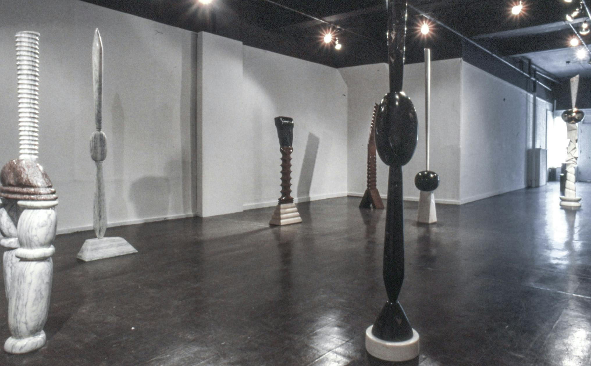 Seven tall sculptures visible in a gallery space. The sculptures are a mix of black, brown and white stone, carved into shapes like ovals, ridged poles, and wavy slabs arranged in different ways.