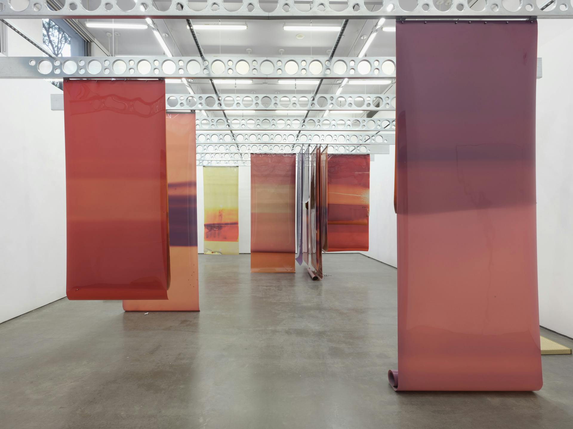 A wide view of long earth tone sheets of film of various colours suspended from industrial steel ceiling beams. 