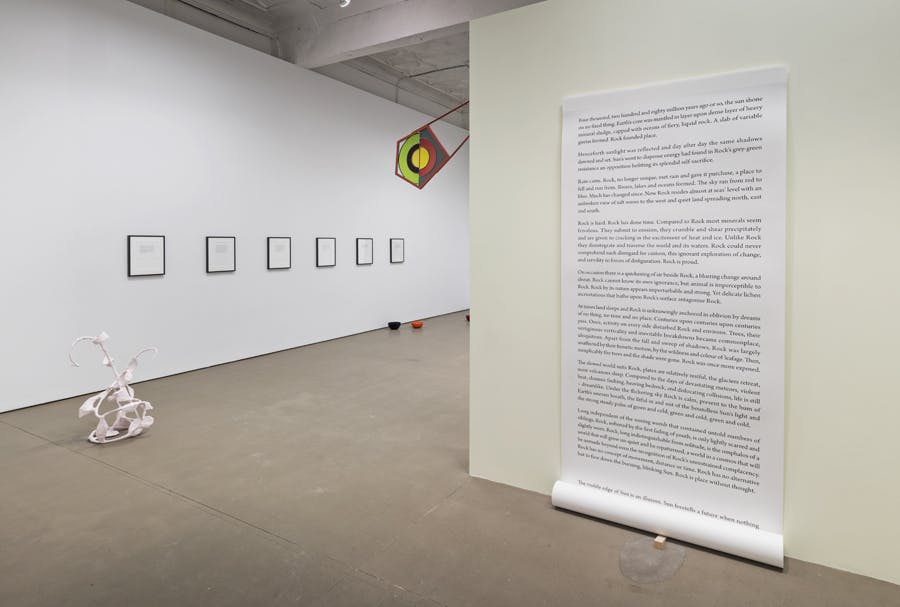 Several artworks are installed in a gallery space. A large scroll paper with some texts is hanging from a wall, and an abstract biomorphic-shaped sculpture sits in front of the other wall.  