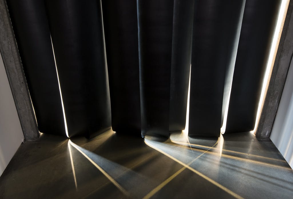 A  black vinyl strip curtain is installed in a hallway at the entrance to a gallery. Rays of light come into the hallway through the narrow gaps between the strips. 
