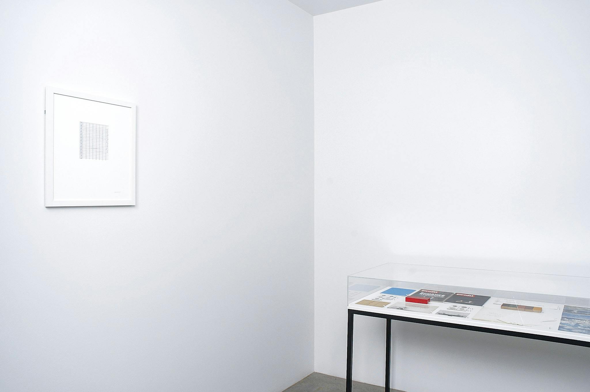 An installation image of artworks by Micah Lexier. A variety of objects such as postcards and magazines are placed in a display case. A drawing that looks like a graph is mounted on a wall.