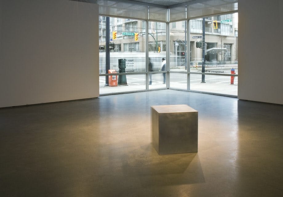 A sand-coloured cube sits in the middle of the gallery space. The cube is slightly shaking. The cube is about toddlers height. The window at the corner of the room shows the outside street scenery. 