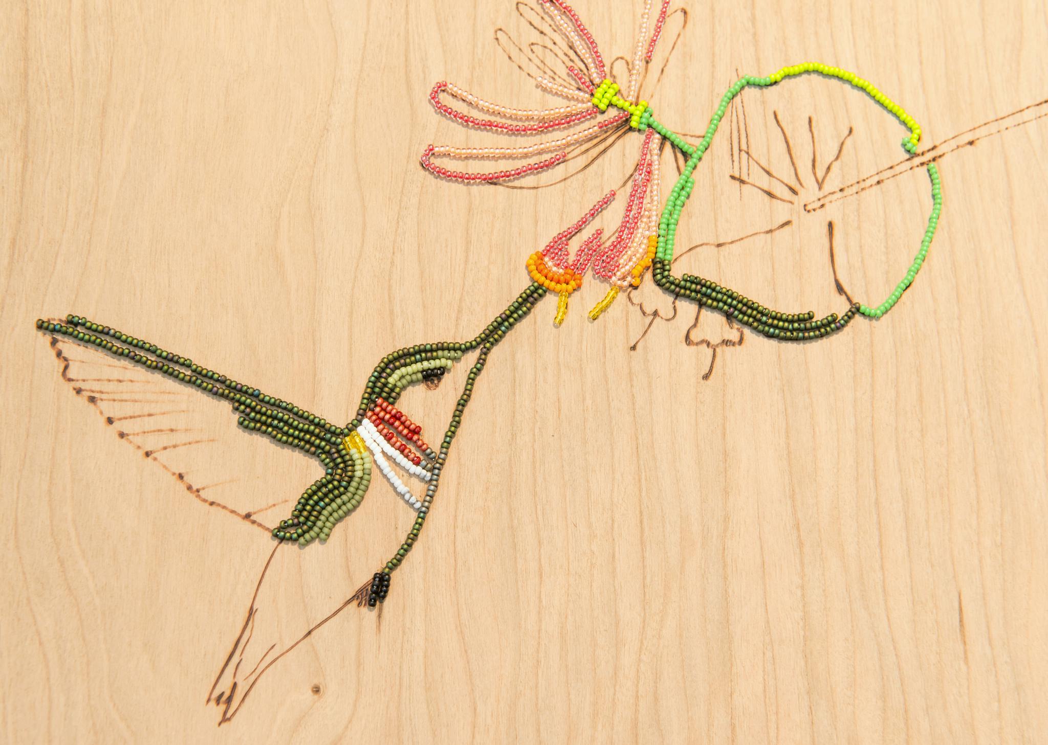 An image detail of a wood veneer panel with a rendering of a hummingbird drinking nectar out of a flower. The rendering is composed of bead-embroidered and wood-burned lines.