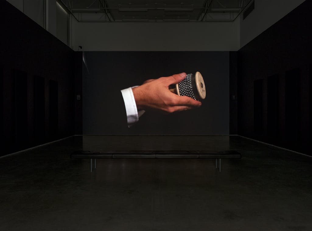 A single-channel video is projected on a screen in a darkened gallery. It depicts hands holding a cabasa instrument.