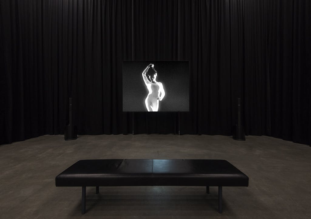 In a gallery enclosed by black curtains, a single-channel video is projected onto a screen, the projector atop a plinth in front. In the video, a person poses, raising their hair above their head. 