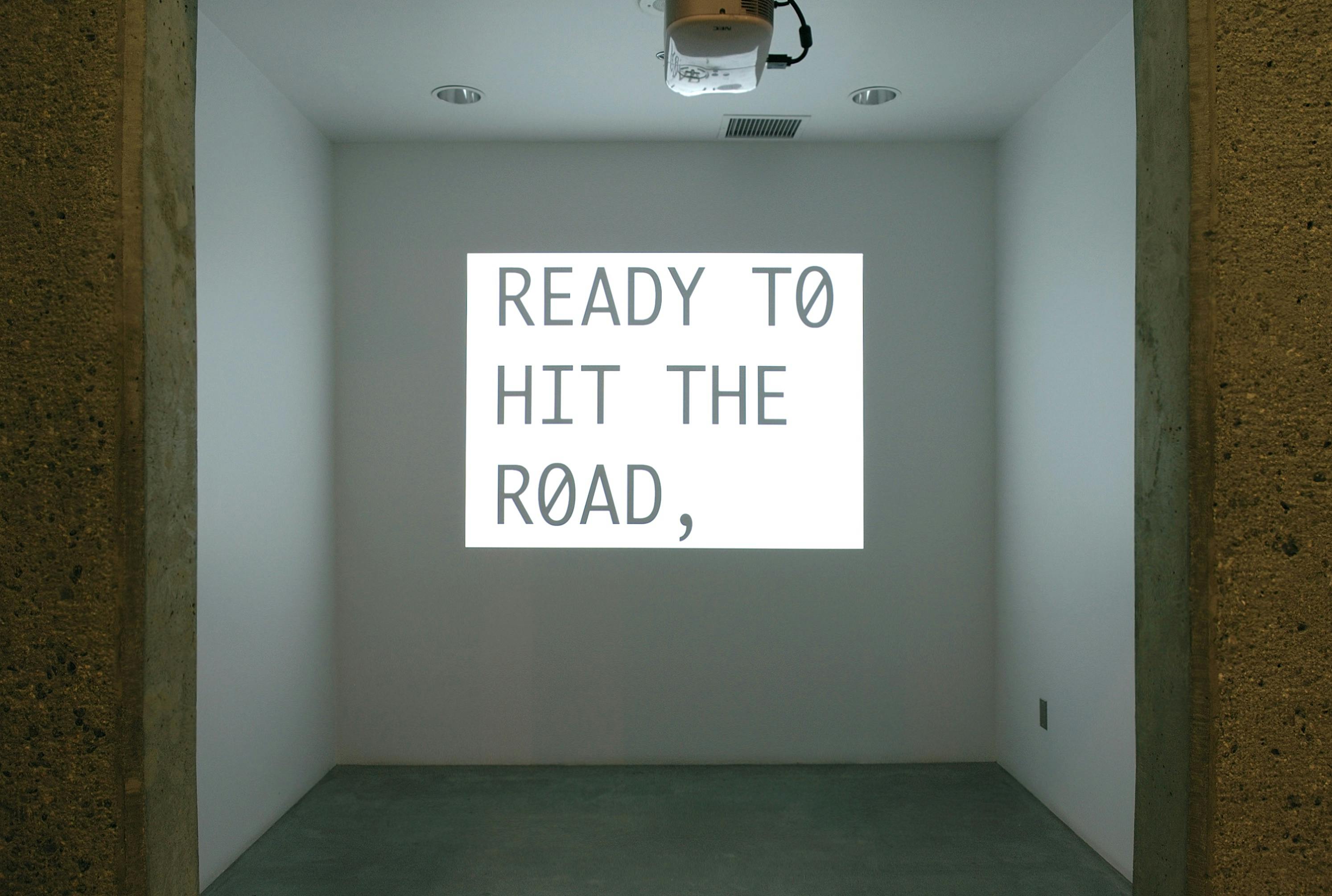 In a small gallery space, a projector is hanging from the ceiling. It projects a slide to the wall. The phrase that appears in the image reads, “ready to hit the road,” in upper letters.