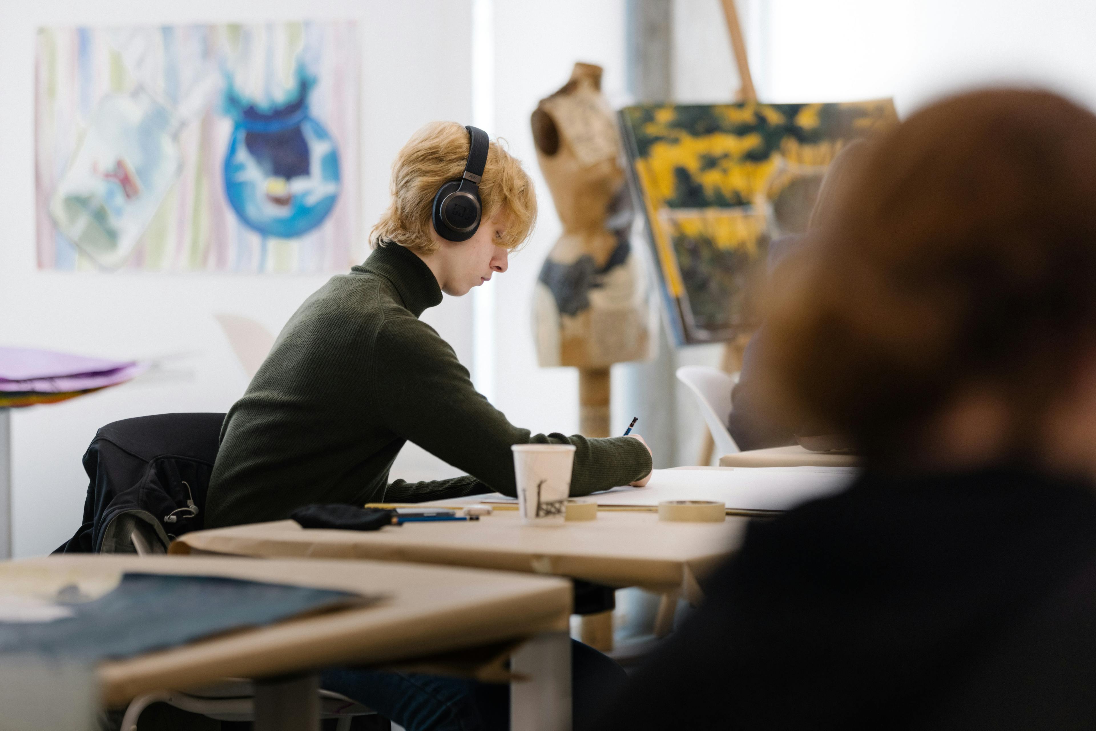 A young artist sits at a desk with headphones on. In the background, paintings and sculptural artworks are displayed.