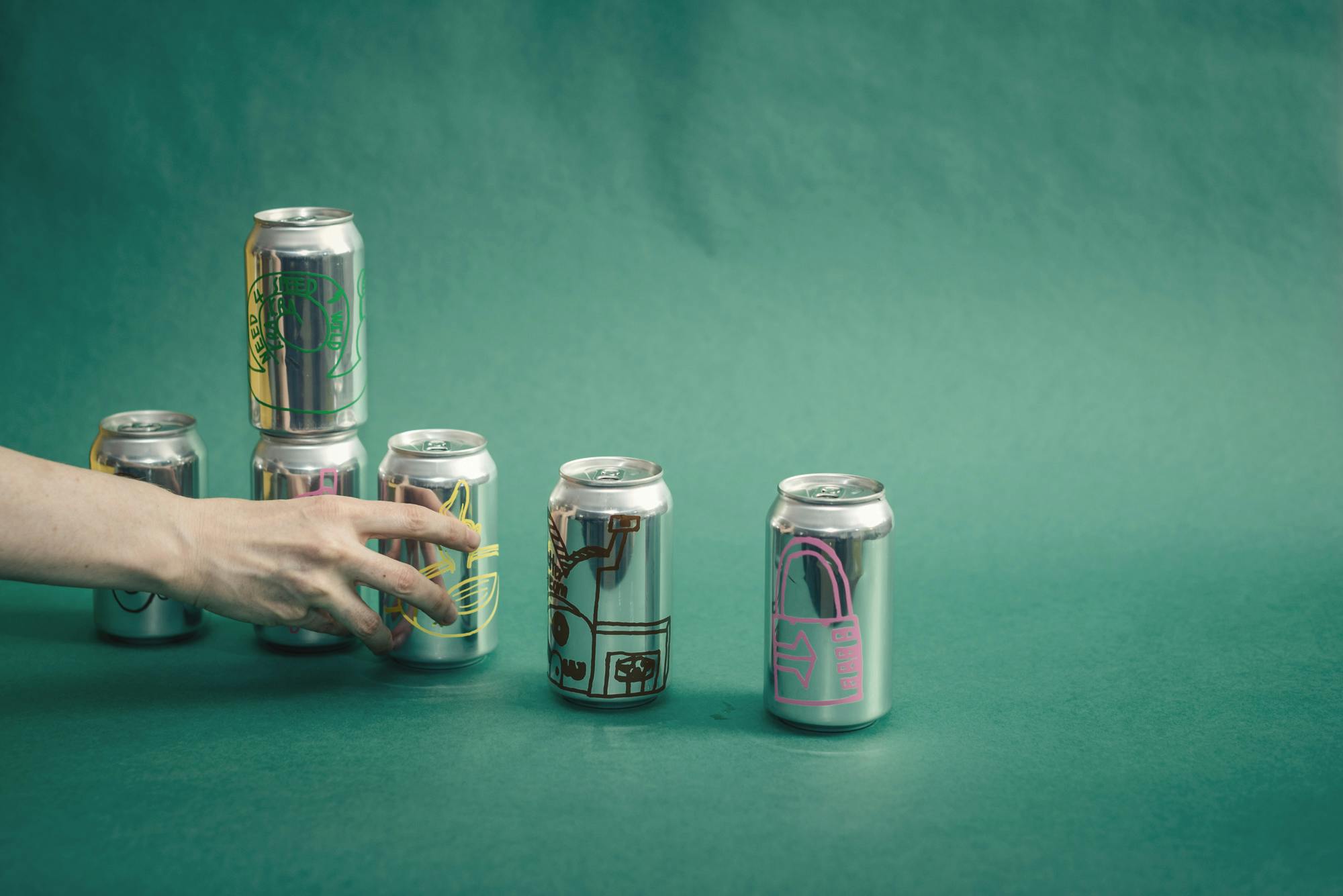 A still image of hands placing cans of soda on a green backdrop. 