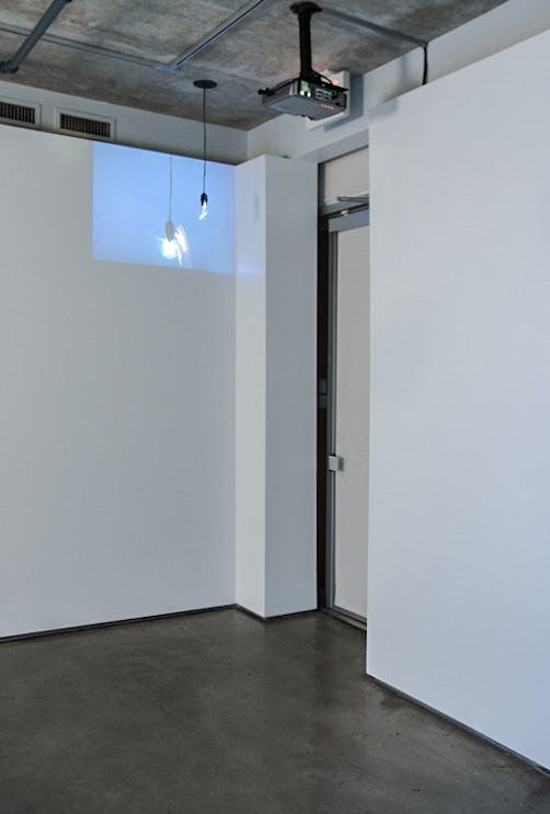 A video projector above the white door projects a moving image on a gallery wall. That video shows a small black object hanging from the ceiling in a blue background. 