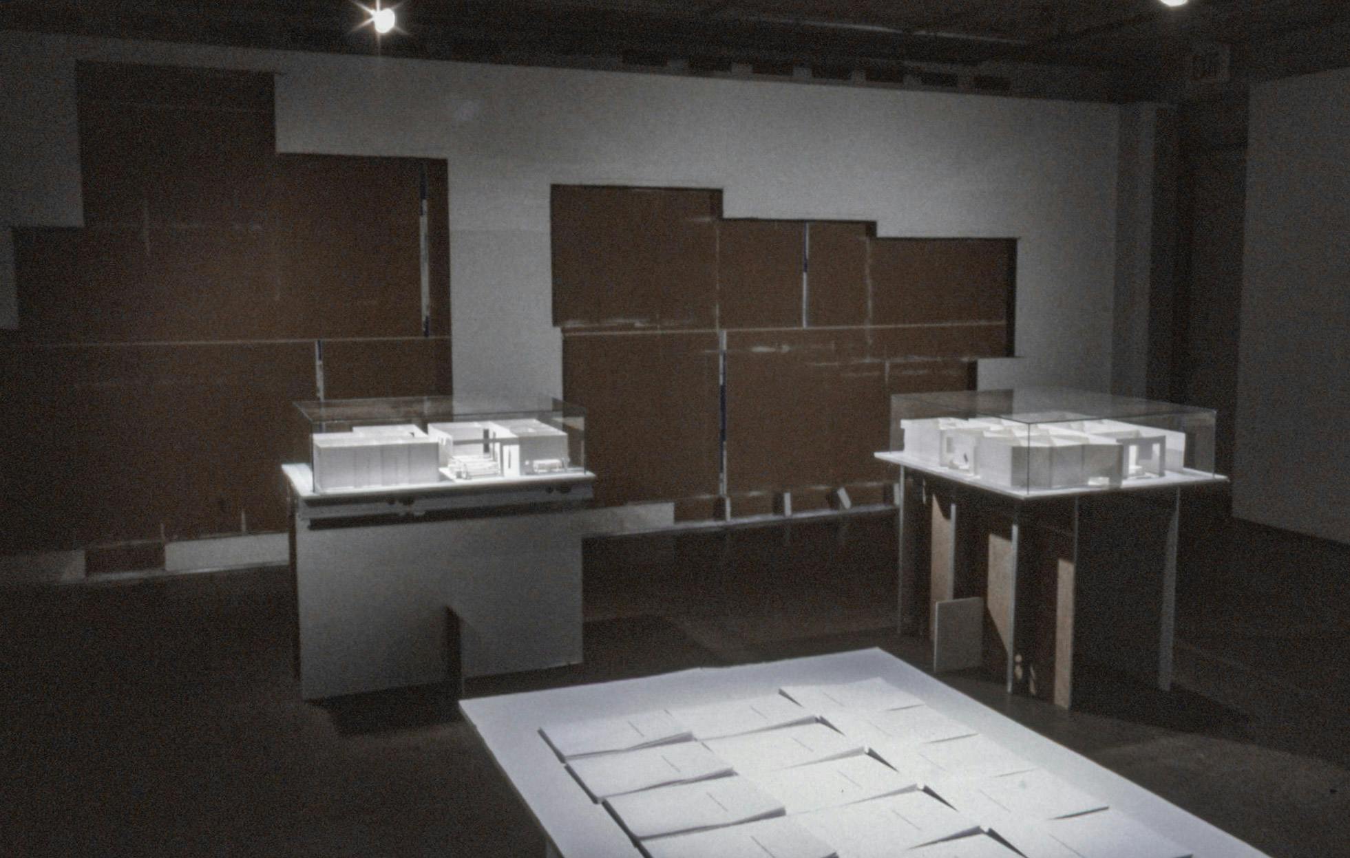 Several white sculptures are installed in a darkened gallery space. Two of them are the mockups of architecture complexes, and the rest is a set of the same documents. 