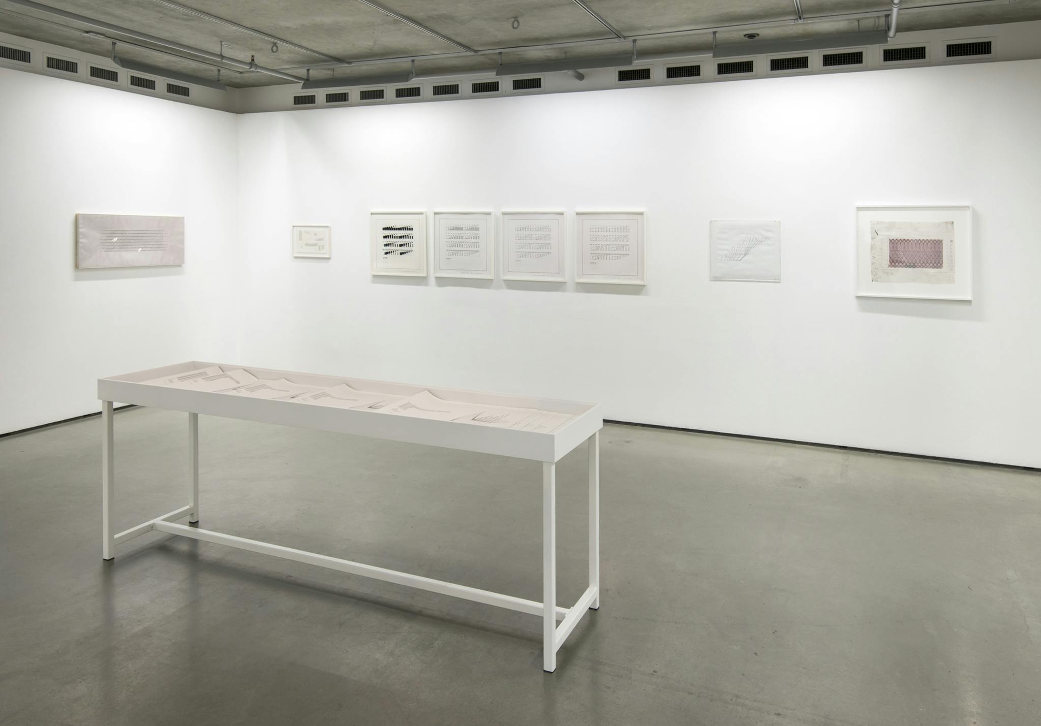 An installation image of Progressions and Rhythms in Eight by Channa Horwitz. Eight framed drawings hang on the white wall of a gallery. A narrow display vitrine with four legs and a glass top sits in front. 