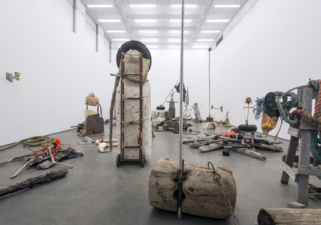 Large sculptures of found materials fill a gallery space. A tall ladder cart holds a huge rock, driftwood branch, and tire. Other sculptures of lumber, driftwood, rope, and metal surround it.