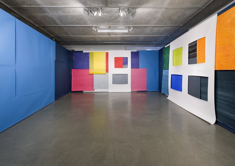 Walls of a gallery are completely covered in layers of paper-like material either cut into colorful squares and rectangles or cut out of large sheets and placed on top.