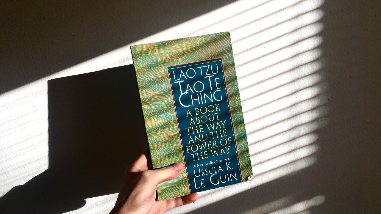 A photograph	 of a hand holding a book titled Lao Tzu: Tao Te Ching: A Book about the Way and the Power of the Way by Ursula K. Le Guin against a white wall. 