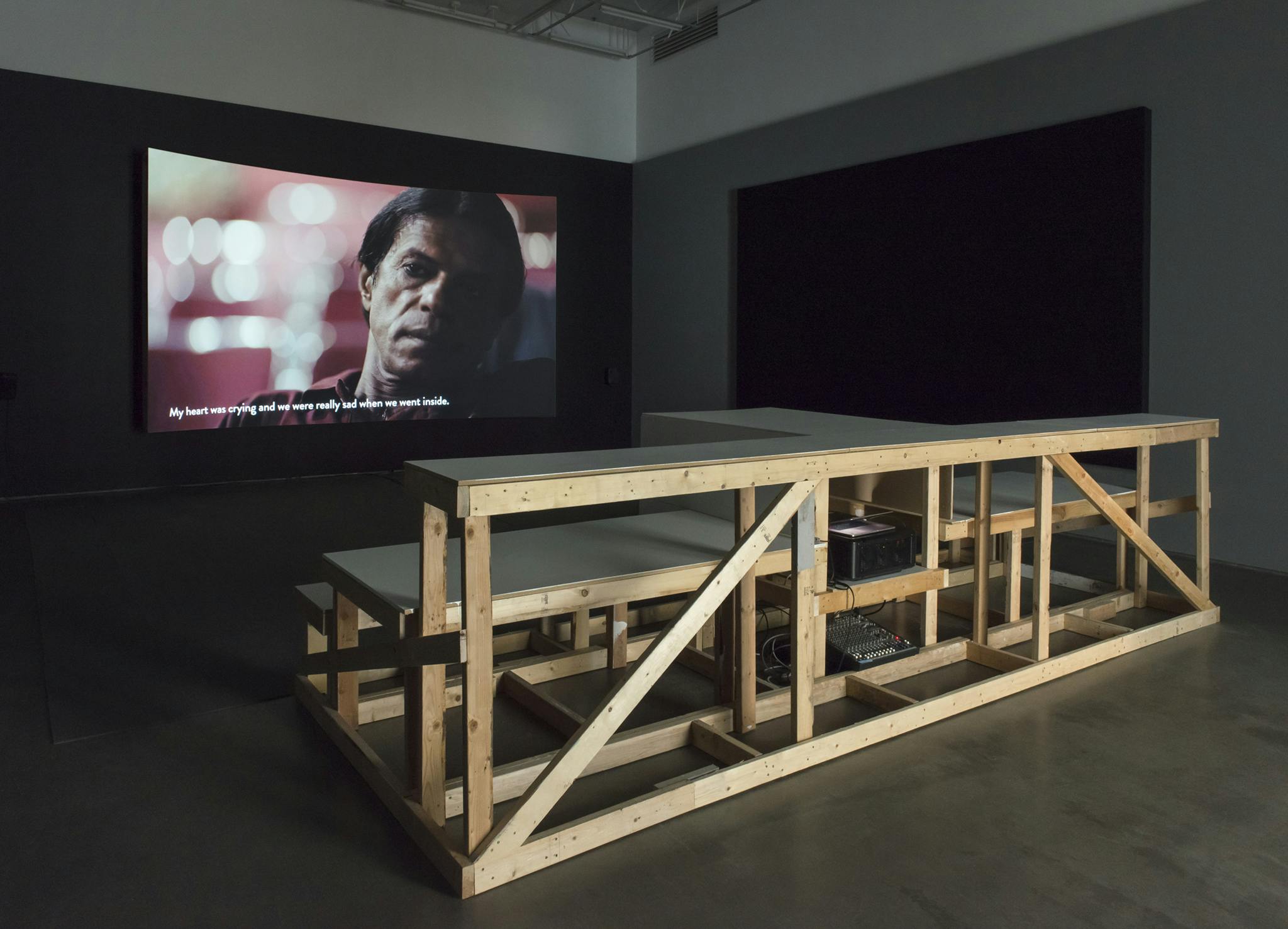 Wooden bleachers pointed towards a large projector screen in a dimly lit gallery. The screen shows a close up image of a person's face with subtitles below.