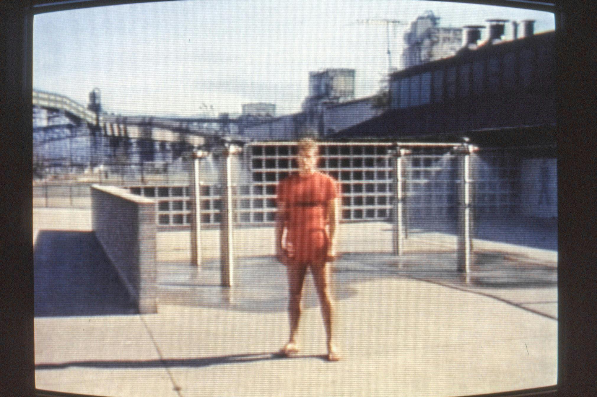 A closeup of a film playing on a small, curved screen. The image shows a person in a red t-shirt and shorts at a spray station in a public park. The image on the screen is grainy from the analog film.