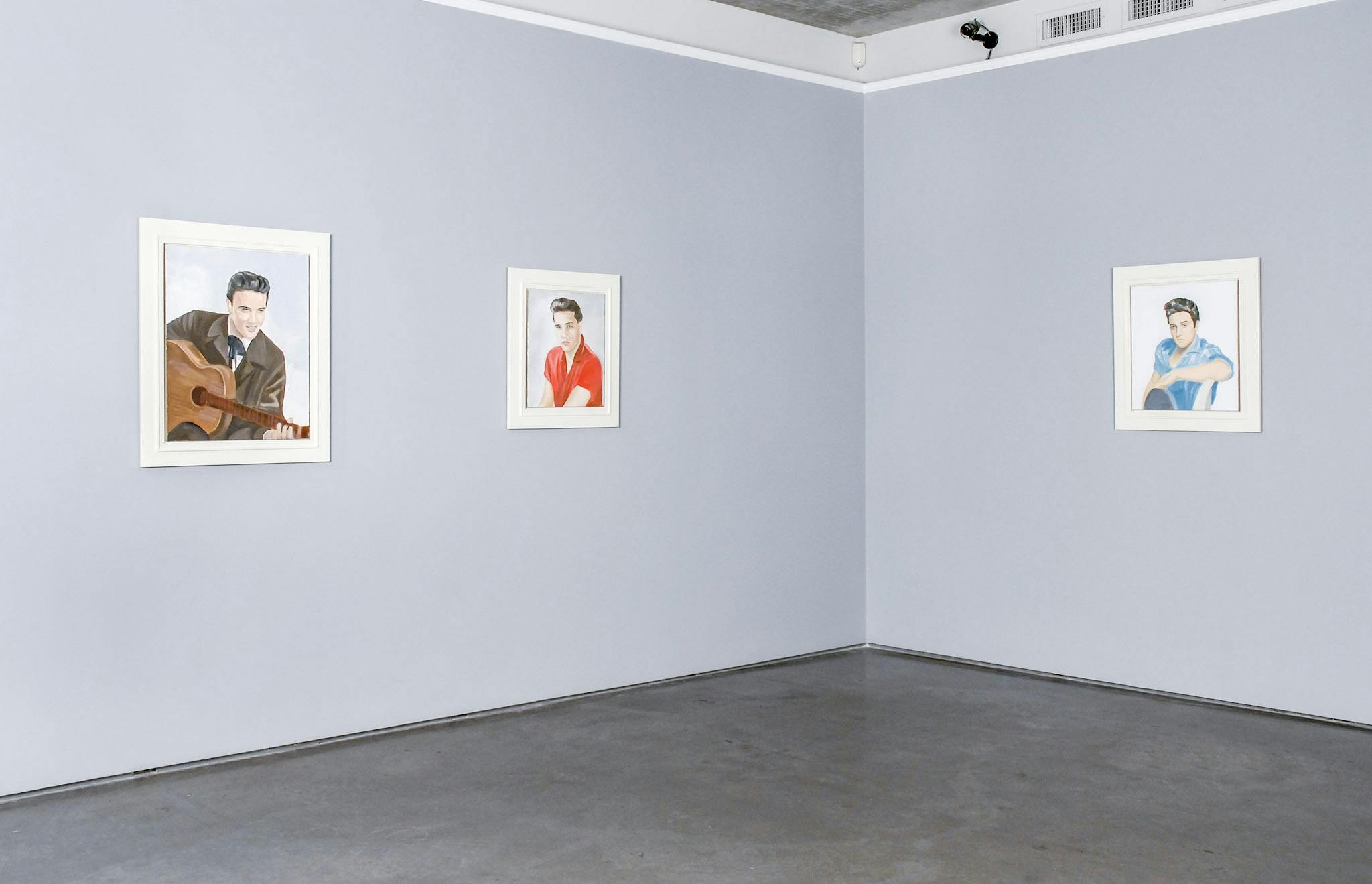Three portrait drawings are mounted on the gallery walls. They are coloured drawings, and each depicted man wears different coloured shirts. One of those depicted men is holding a guitar. 