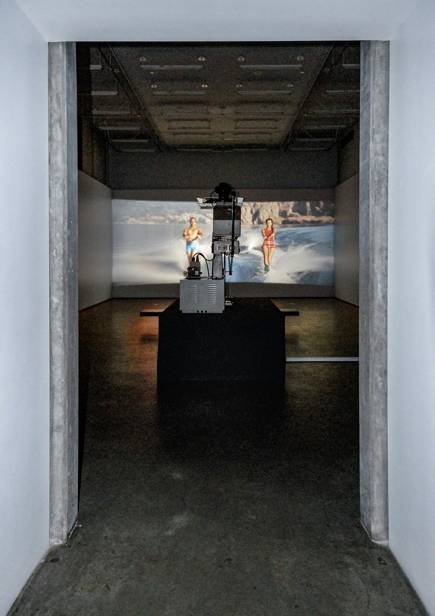 A film is projected on the wall in a gallery. The film depicts a man and woman wakeboarding. A film projector placed on the floor close to the gallery entrance projects this film to the wall. 