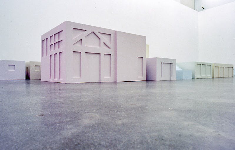 This is a low-angle shot of the installation in a gallery space. Many pale-coloured cube sculptures are placed on the floor. A shape of a house front is carved on one of the  sculptures’ facade.  