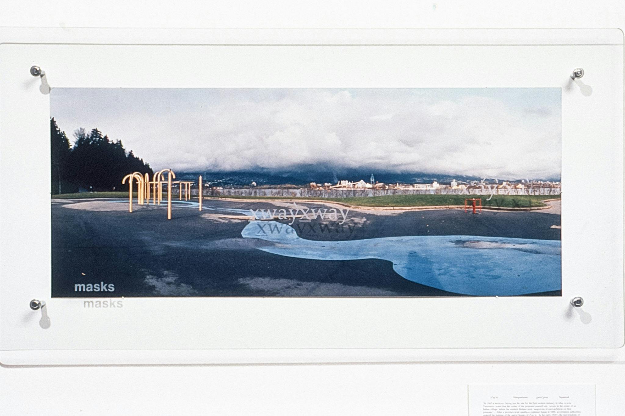 A panoramic photo of a small park mounted on a white wall between 2 panes of glass. The pane on top is engraved with the word "Xcwáy̓x̱waya," the name of a Squamish Village in Stanley Park.