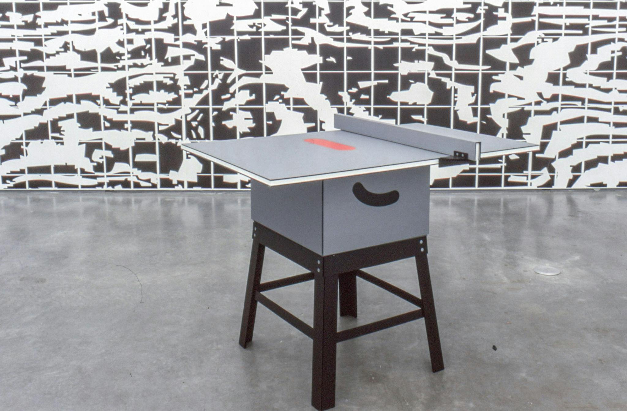 A small grey table is placed in a gallery. Its tabletop looks like that of a ping pong table. The gallery wall is covered with a black and white pattern similar to a wood surface. 