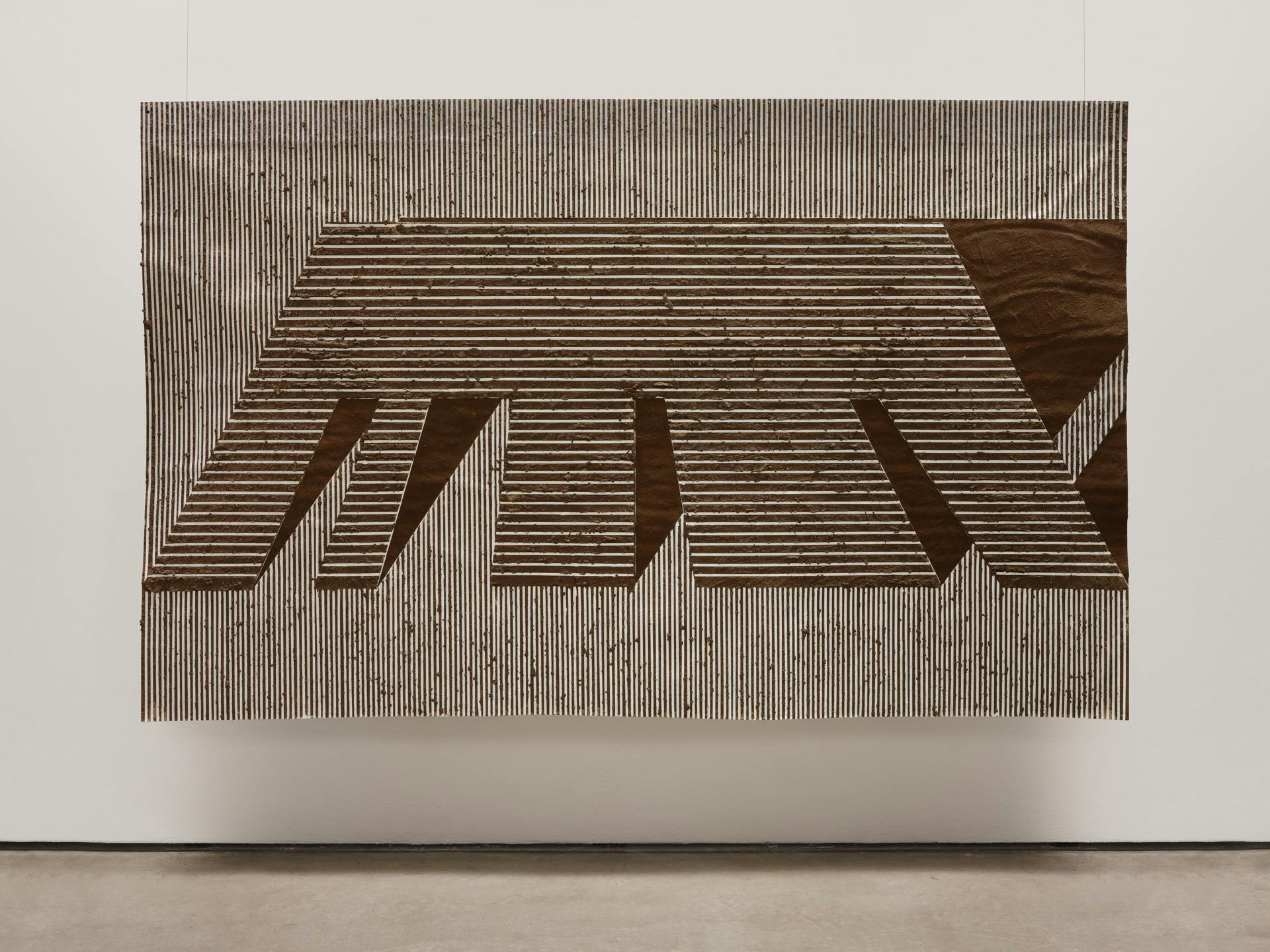 One large scale drawing is suspended against the wall of a gallery space. The drawing of an architectural form is made with striations of brown adobe mud on white paper. 