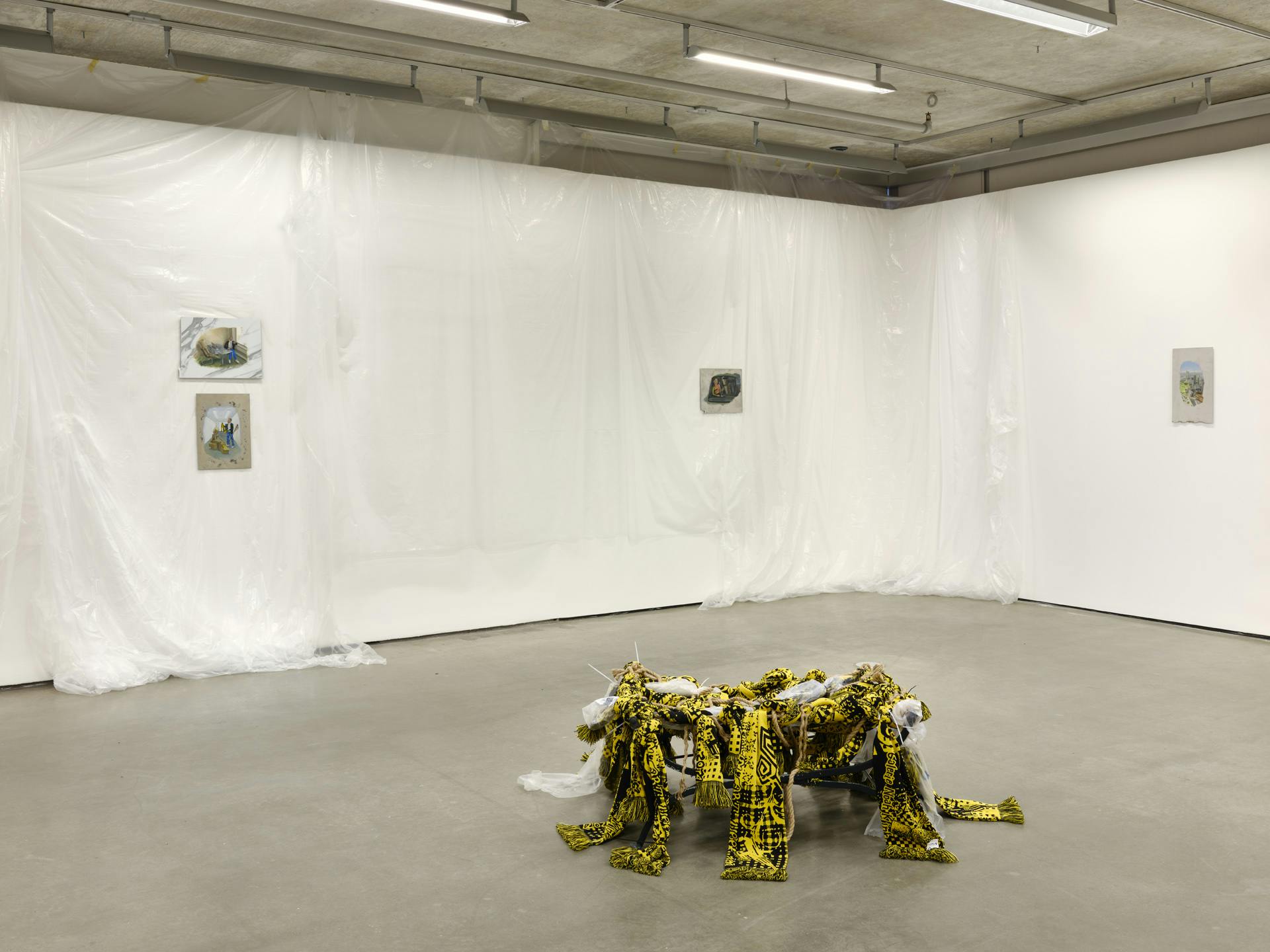 Four small paintings on faux-marble tiles or metal sheets hang on gallery walls draped with translucent plastic sheets. A sculpture made of yellow textiles, plastic and rope sits on the floor. 