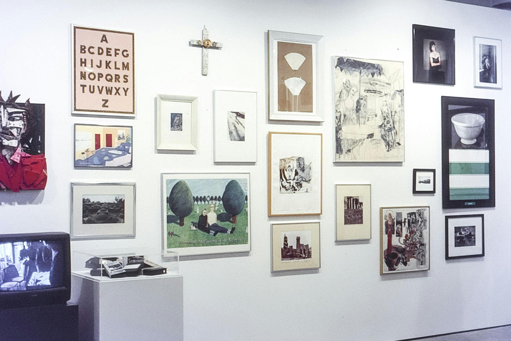 A photo of various artworks mounted on the gallery wall. A white small cross is at the top and placed beneath are paintings and photographs depicting human figures and landscapes in different styles.