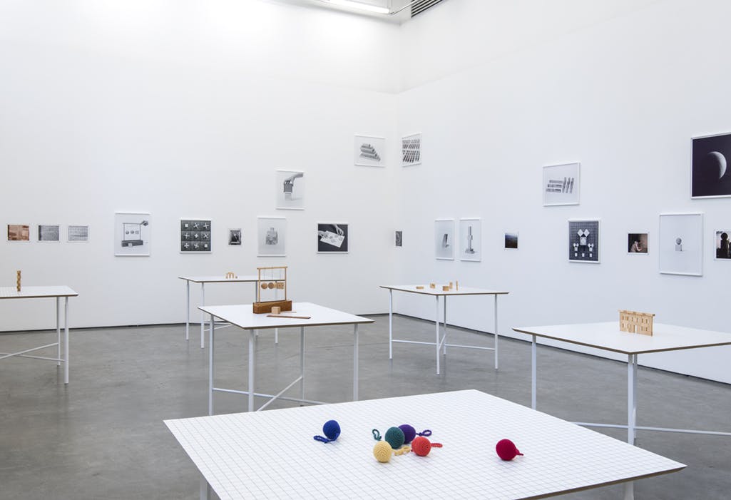 Six objects on white tables in a gallery space. One table holds a group of colourful balls made of wool. Other objects on tables resemble wooden toys. Black and white photographs hang on the walls. 