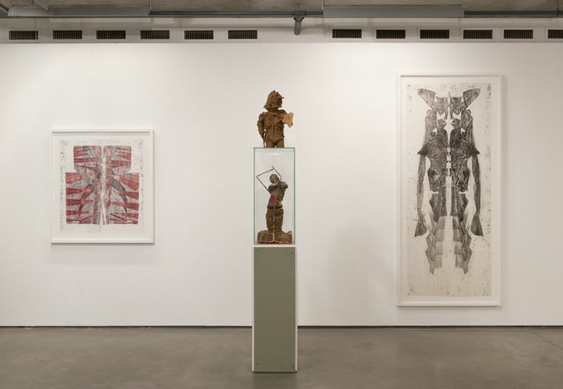 Two human form sculptures and two framed works installed in a gallery. The sculptures appear rusted with copper-coloured surfaces and stand on a plinth.