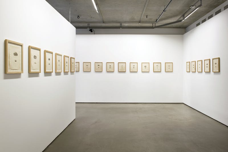 An installation view of a gallery with multiple framed works mounted on three walls. Each small-sized print has one larger and one smaller image, vertically composed with the larger above the smaller.