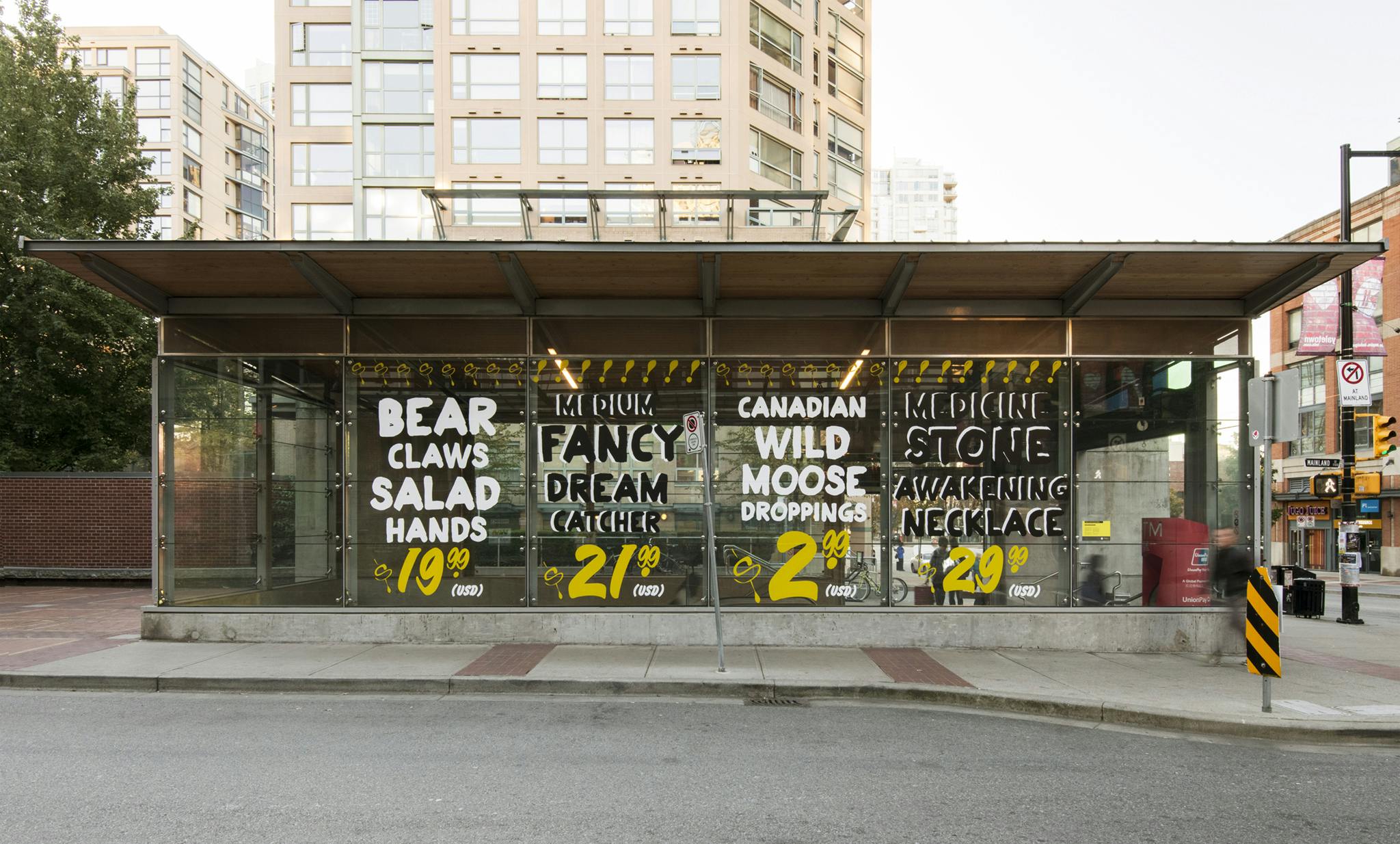 Text-based artworks in vinyl are installed on the glass facade of Yaletown-Roundhouse Station, resembling hand painted signs. The letters are all capitalized in black, yellow, or white. 
