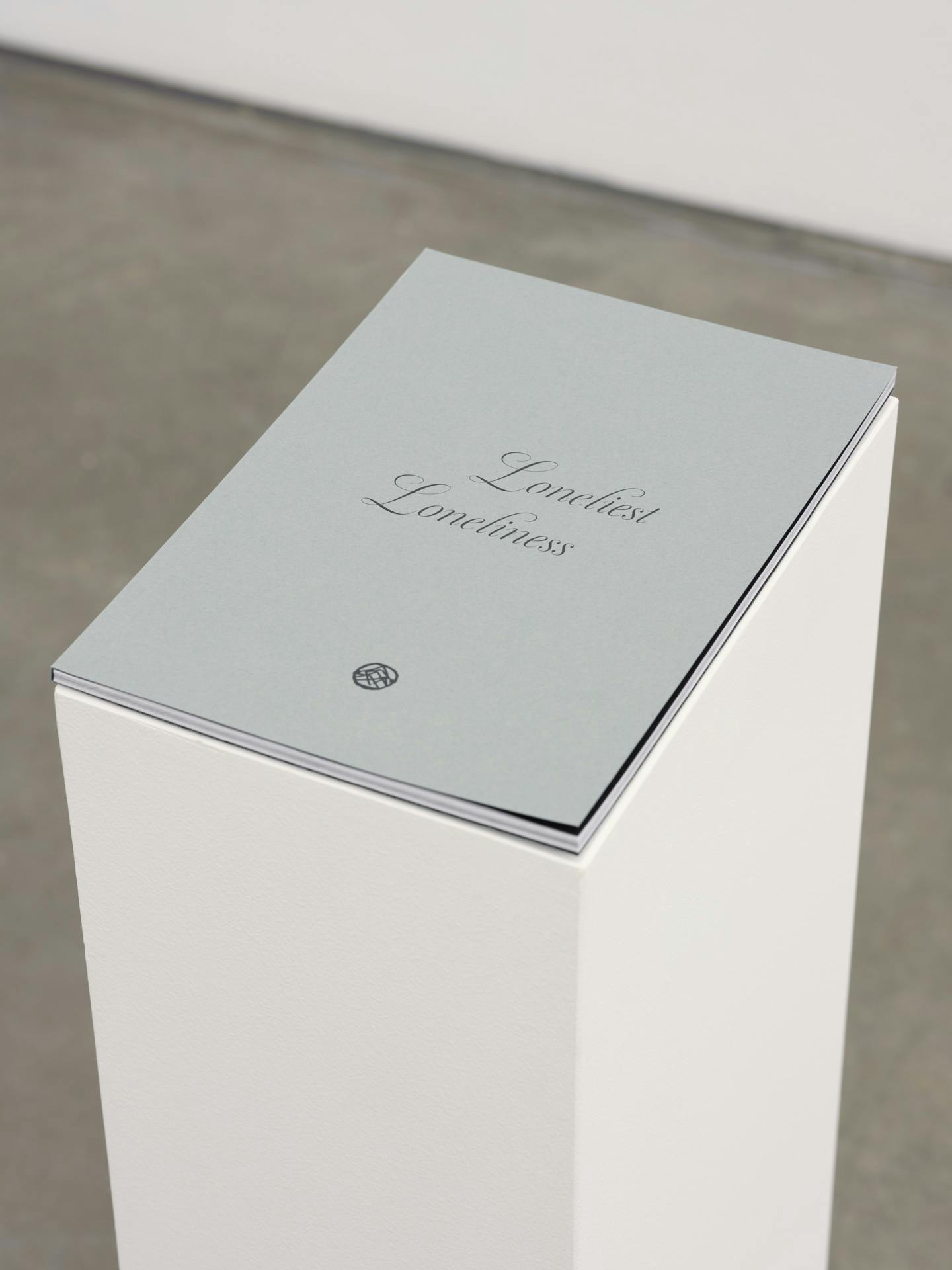 A thin artist book by Kathy Slade lying closed on a white plinth, with the title Loneliest Loneliness visible on the cover.