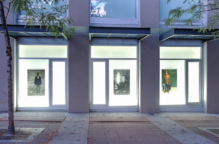 An installation image of a work by Daniel Olson in CAG’s window spaces facing a sidewalk. Three poster-shaped photography works are mounted on an illuminated wall behind glass windows. 