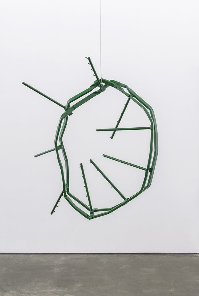 A steel sculpture hangs from a gallery ceiling. The sculpture is painted green, its form two concentric circles. The circles are misshapen and bent, with metal rods protruding outwards from one.