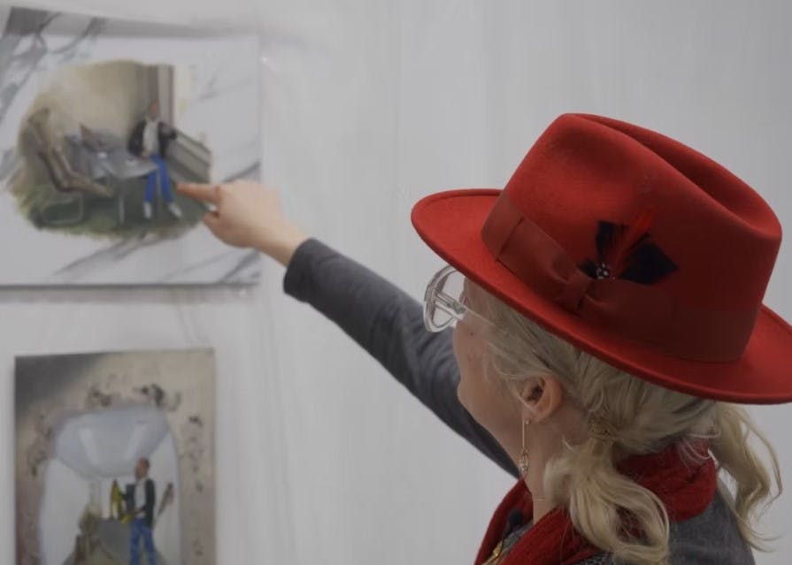 A still image of psychic Star Magic pointing the finger at one of Alison Yip's works on the wall. Star Magic wears a red hat and glasses. 