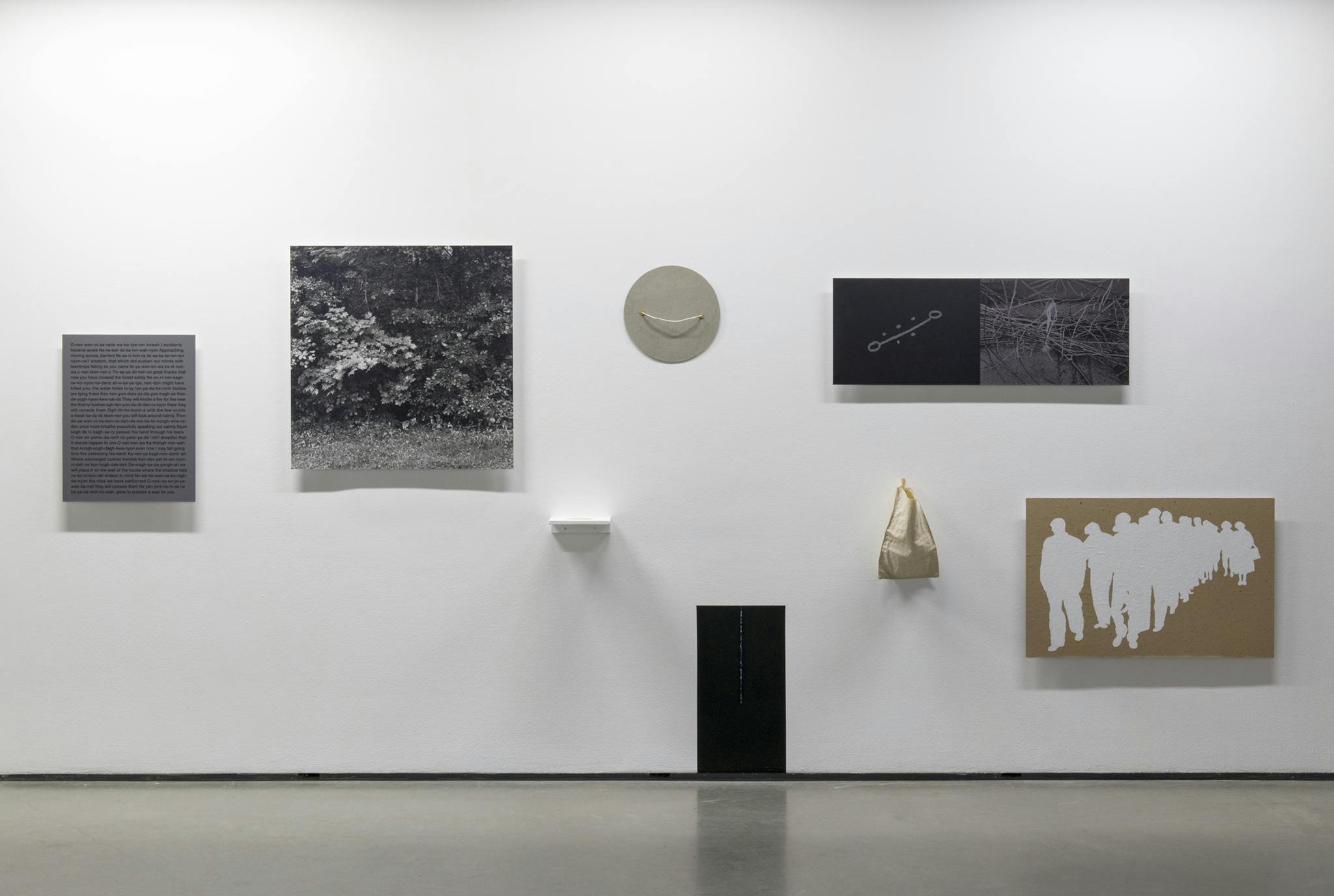 Multimedia objects hang on a gallery wall. Each object differs in form, from a text-based print, a black and white photograph, to a very small shelf and a cloth bag.
