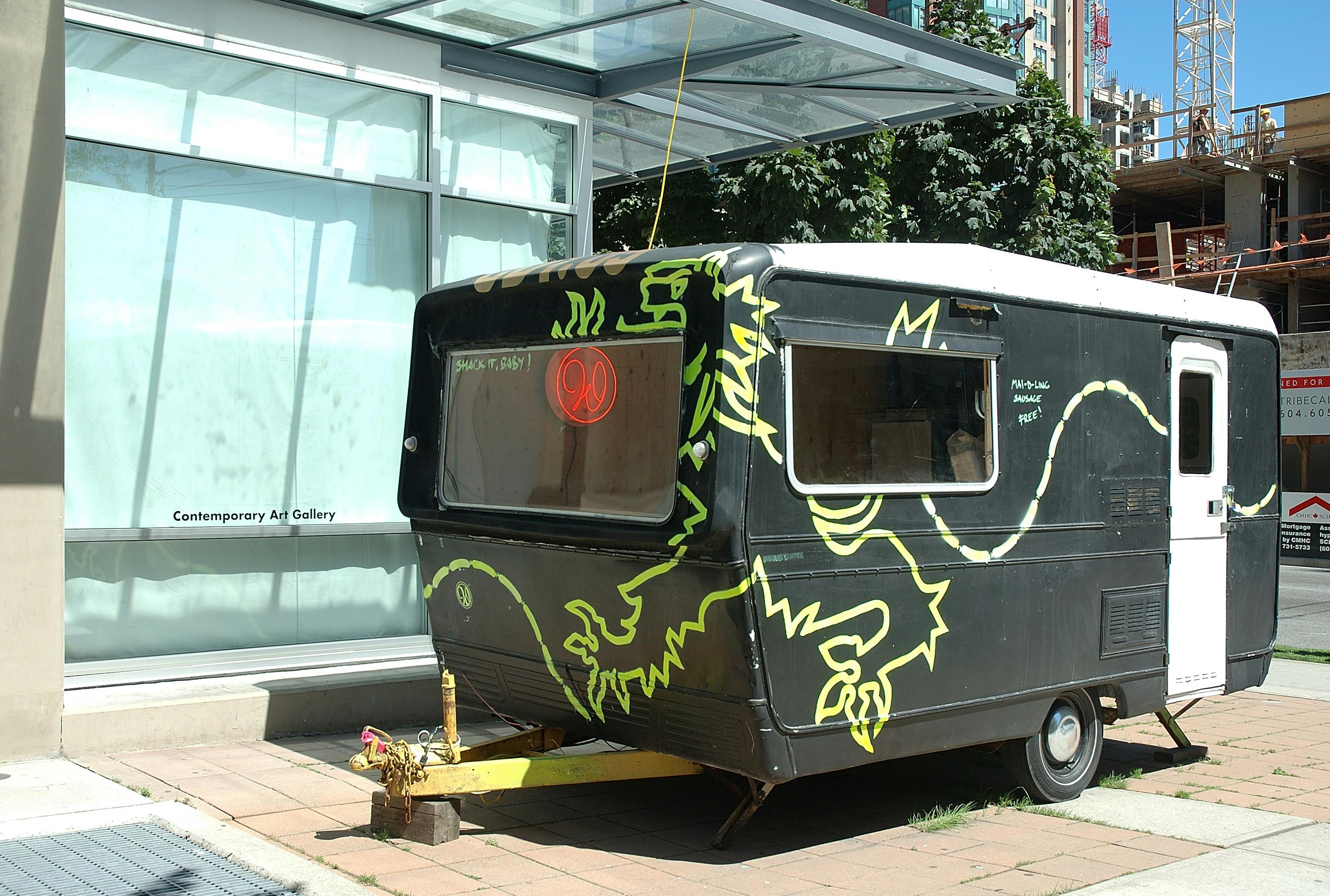 An installation image of a black camping car placed right outside the gallery. A yellow lion or dragon shape is painted on the facade of the car. 