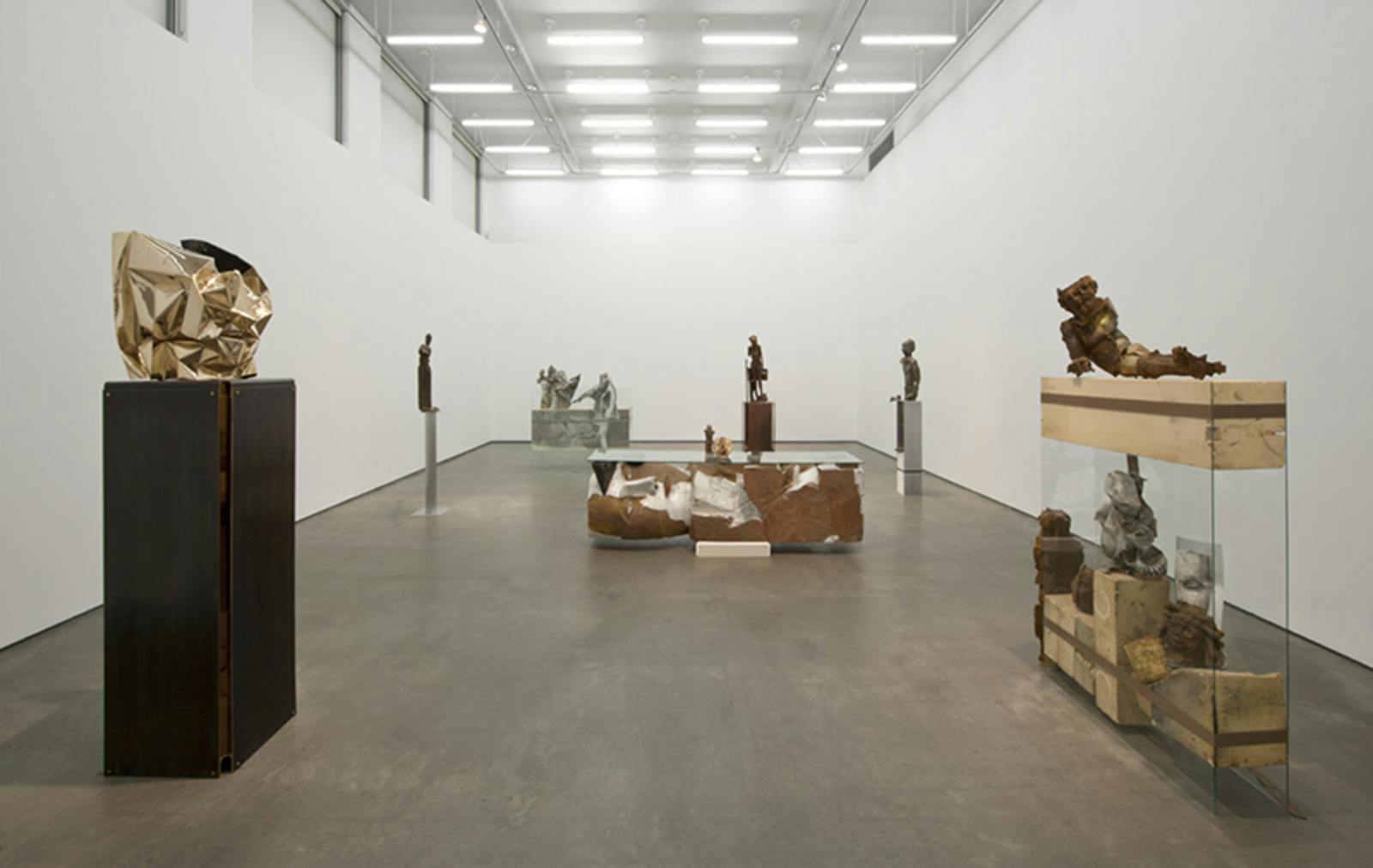 Multiple sculptures on plinths of different heights and materials and design are installed in a gallery space. One has an abstract form with a metallic surface while the rest resemble human forms. 