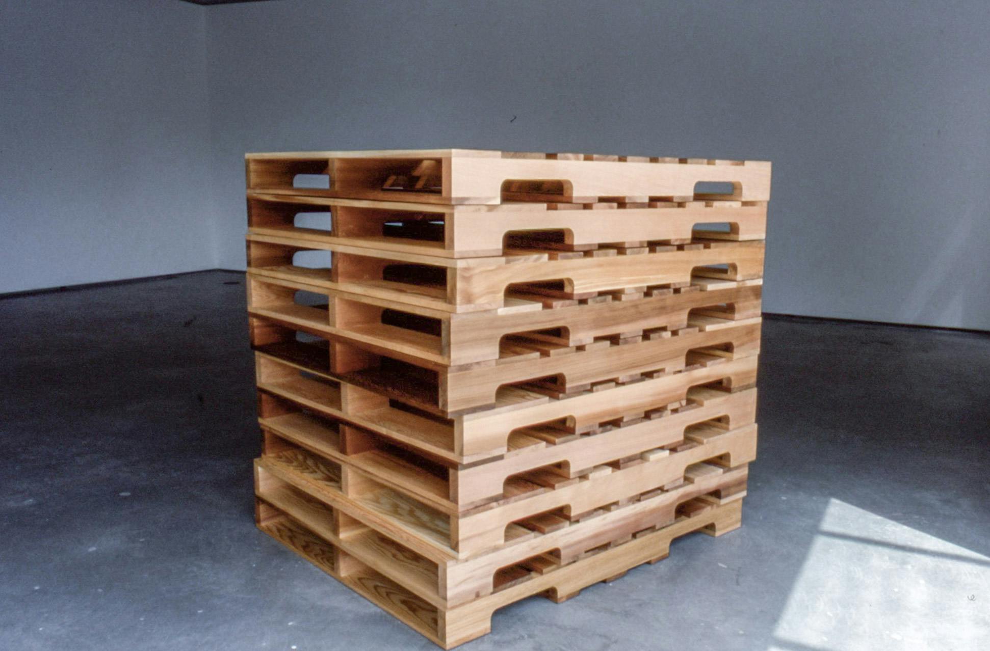 A stack of ten wood pieces, which look like large duckboards, is installed in the middle of a gallery space. They are identical in shape and size. None of them are painted. 