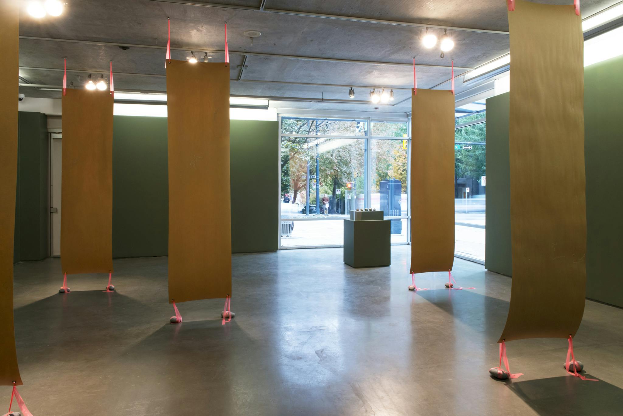 Installation shot of five panels of veneer stretched vertically from the gallery floor to ceiling. A green vitrine sits in the background, placed in front of a large corner window.