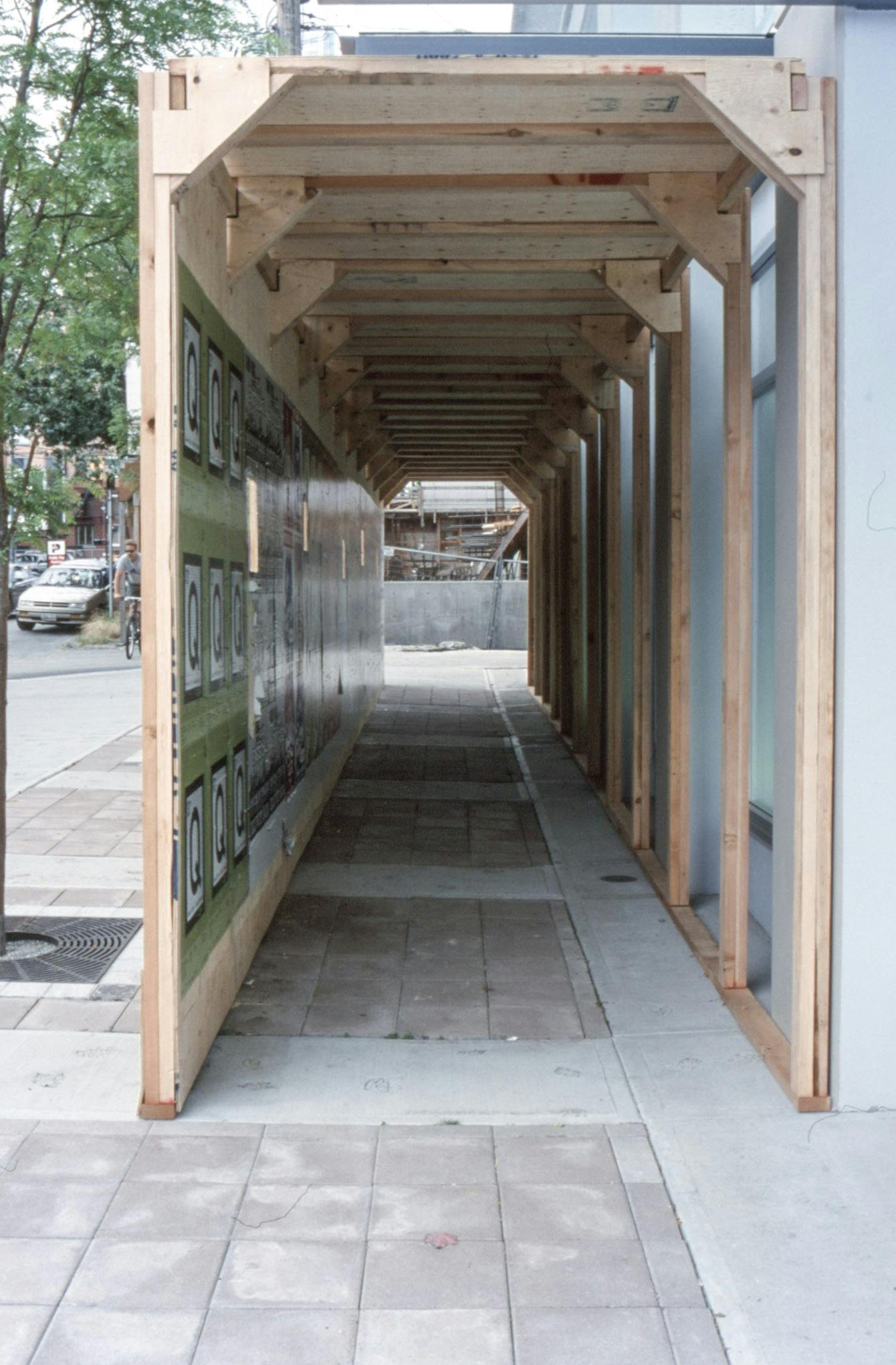 An unpainted wooden tunnel is built in front of CAG’s window spaces. It resembles construction hoardings. The title of this work is “Unlimited Growth Increases The Divide.”