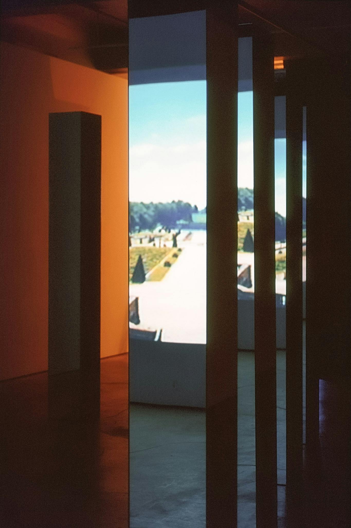 A close-up view of the mirrored columns installed in the gallery. They stand on the floor and reflect a photographed image of a garden that is projected on the wall. 