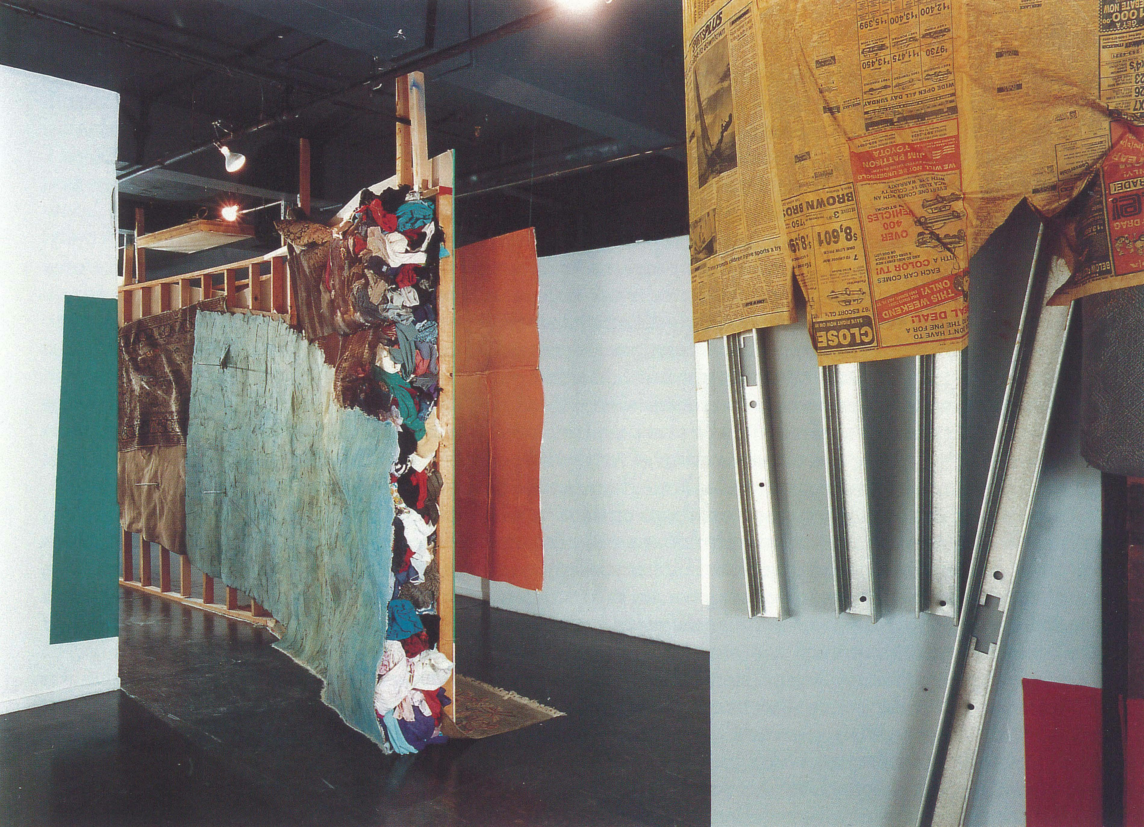 In a gallery space, several large-sized sculptures are installed in a gallery space. Large dyed flat sheets of paper, newspapers, fabrics, and metal frames are attached to the walls. 
